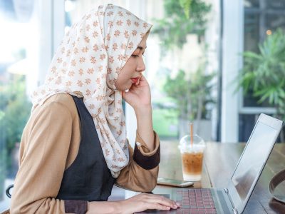 Young Thai Melayu female freelancer wearing hijab work using a laptop on wooden table in coffee shop. Modern woman life style.