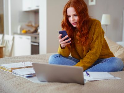 Attractive Young Woman with Red Hair Working from Home - Female Entrepreneur Sitting on Bed with Laptop Computer, Paperwork and Checking Cell Phone from Comfort of Home