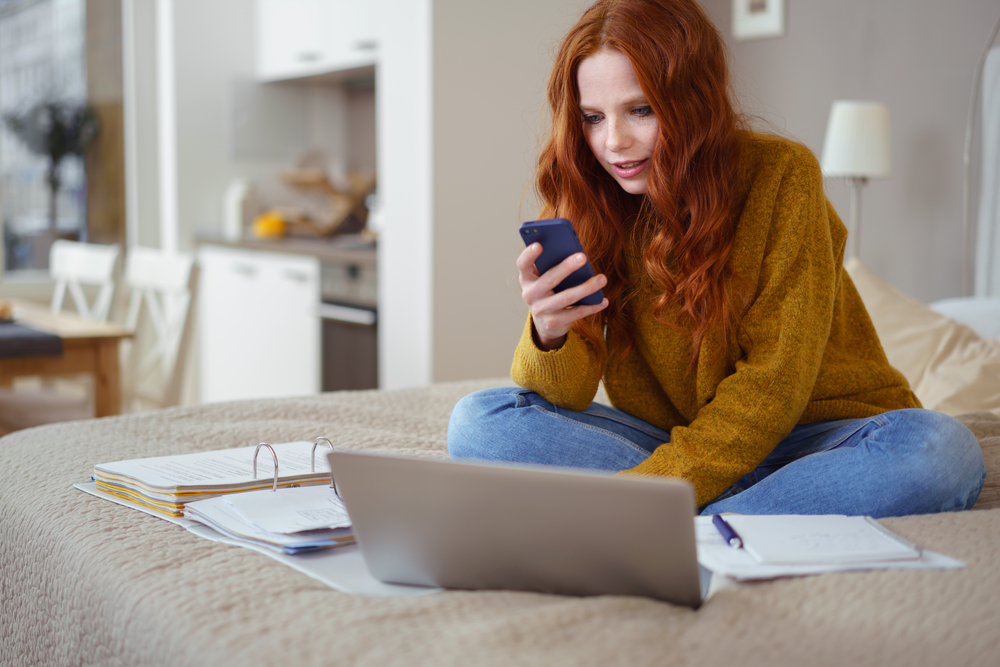 Attractive Young Woman with Red Hair Working from Home - Female Entrepreneur Sitting on Bed with Laptop Computer, Paperwork and Checking Cell Phone from Comfort of Home, job hunt