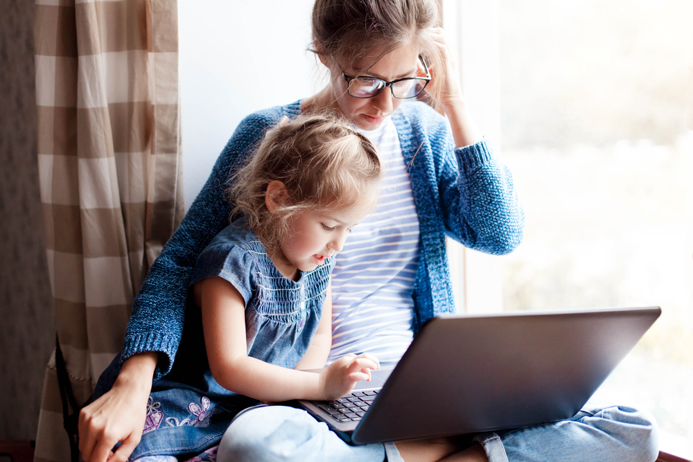 Working mom works from home office with kid. Mother and daughter read news. Woman and cute child sitting on window sill.