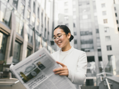 Young attractive Asian female banker or accountant in glasses is reading newspaper outside a modern office center or a bank