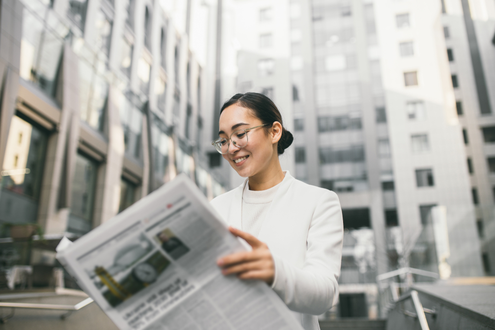Young attractive Asian female banker or accountant in glasses is reading newspaper outside a modern office center or a bank