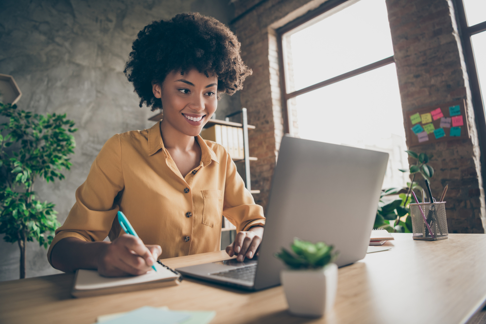 Photo of cheerful joyful mixed-race woman in yellow shirt smiling toothily writing down notes holding training for students to be executives at laptop desktop table, flexible working