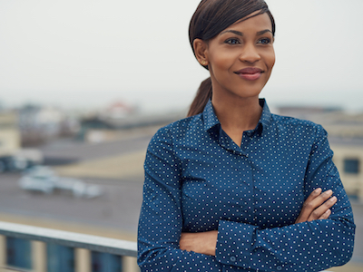 Confident friendly black business woman standing with folded arms on the rooftop of an urban commercial building smiling as she looks to the side of the camera, self-acceptance