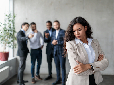 Workplace Sexism And Bullying. Unhappy Victimized Businesswoman Standing While Her Male Colleagues Whispering Behind Her Back Standing In Modern Office