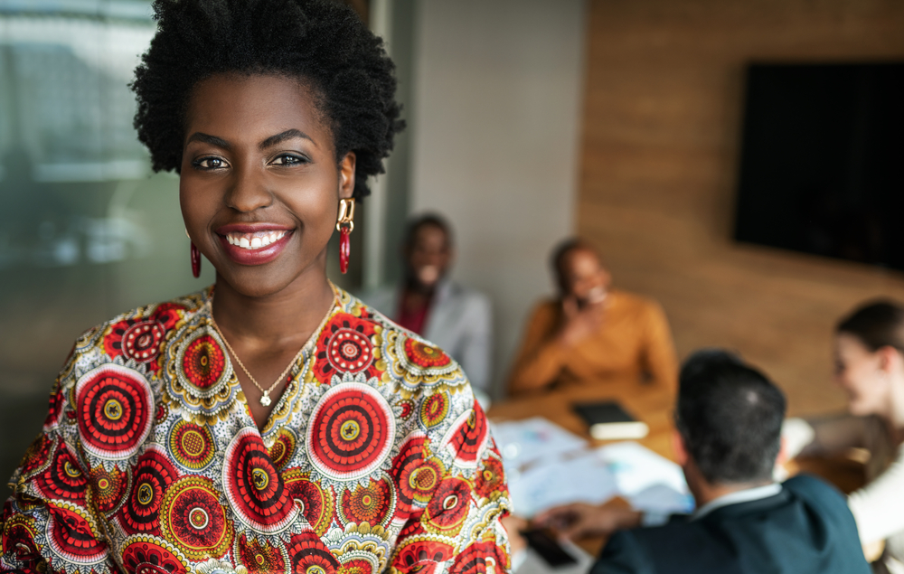 close up of beautiful young smiling professional black african business woman, coworkers hold a meeting in background, black female founders list
