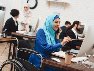 Disabled arab woman in hijab in wheelchair working in office. Woman is working on desktop computer and talking on phone.