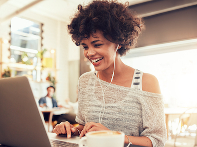 Image of happy woman using laptop while sitting at cafe. Young african american woman sitting in a coffee shop and working on laptop, workplace wellbeing