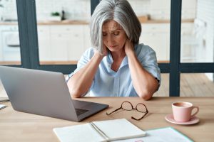 Tired stressed old mature business woman suffering from neckpain working from home office sitting at table. Overworked senior middle aged lady massaging neck feeling hurt pain from incorrect posture. Menopause