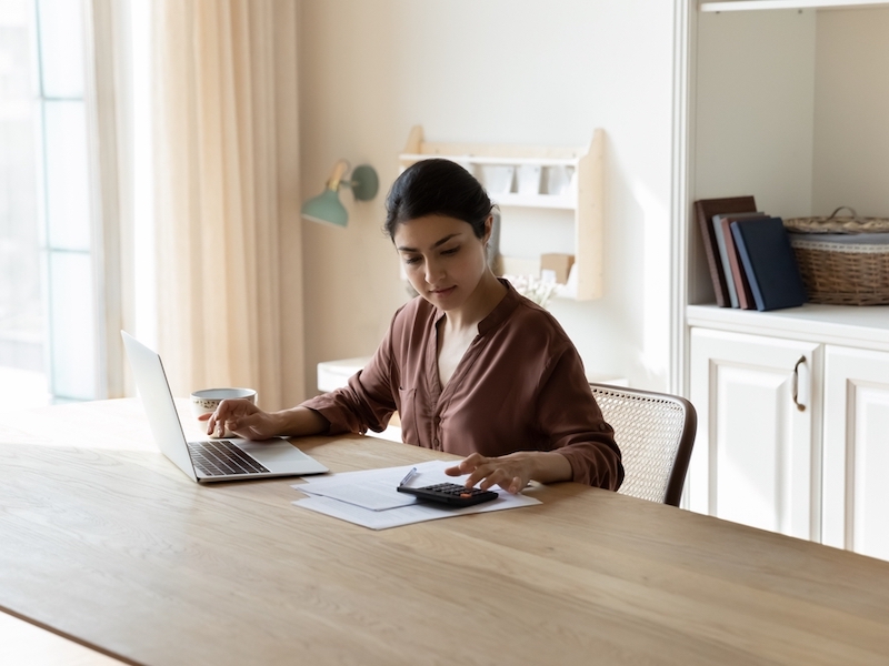 Woman sat at table busy in personal expenses management, use calculator calculate household utilities, pay bills through secure e-banking app on laptop. Finances, accounting concept