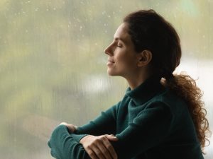 Side shot of serene woman relaxed by window with closed eyes listen sound of raindrops feel pleasure. Calm young lady enjoy breathing fresh ozonized air after rainstorm.