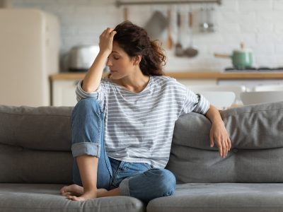 Exhausted frustrated young woman touching forehead, sitting on couch alone, suffering from strong headache, hormones or migraine, worried girl thinking about problems, divorce or break up, lost in thoughts