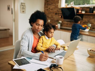 Black working mother taking notes while daughter is sitting on her lap and using laptop at home. Small boy is in the background.